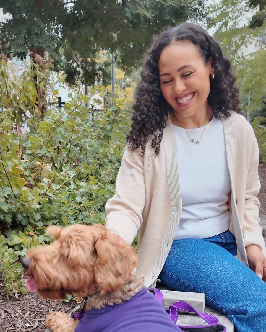 A woman in a cream cardigan smiling on a bench beside her dog, who wears a purple sweater