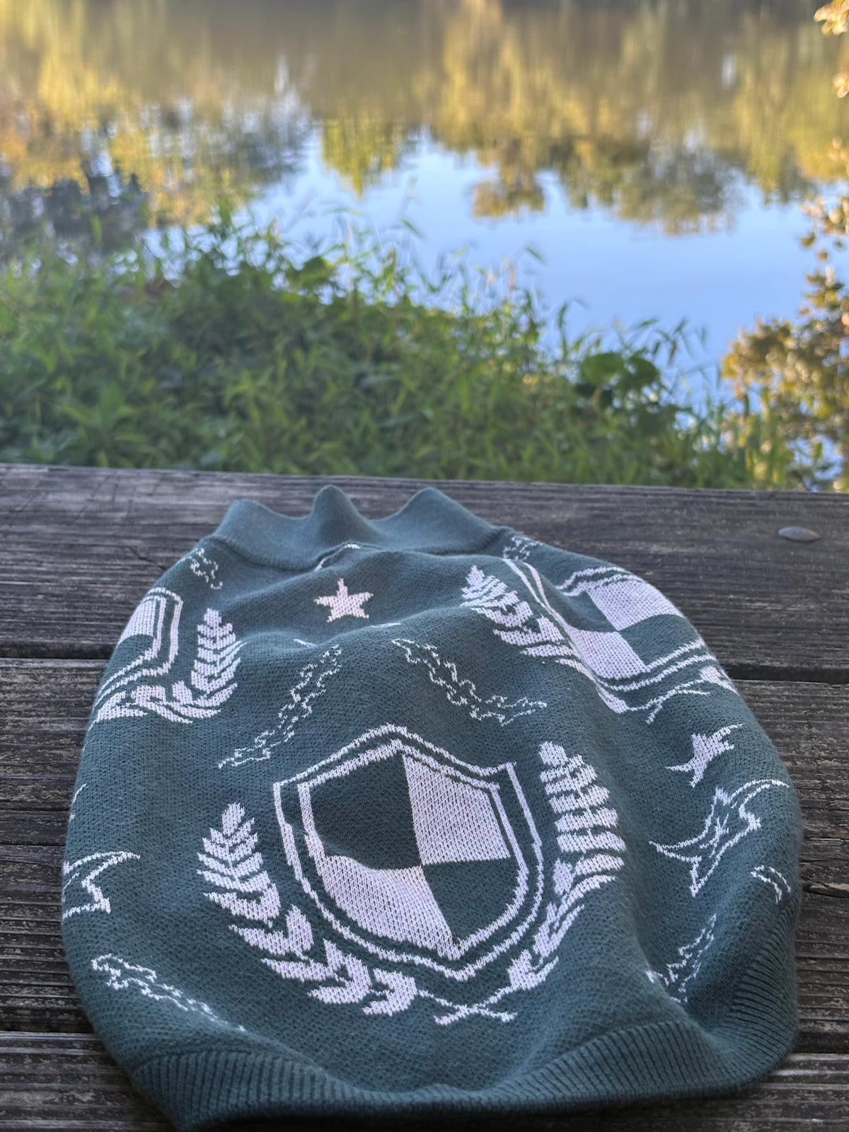 Green crest dog sweater with a cream shield and laurel pattern, laid flat by the lake